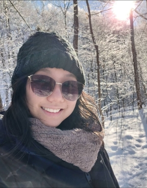 Stefane is an Asian woman wearing a knitted hat and scarf and sunglasses, enjoying the sunshine in a snowy forest.