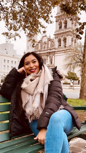 Arani is a woman with dark hair wearing lipstick and a warm jacket and sitting outside on a bench with the facade of a baroque church in the background.