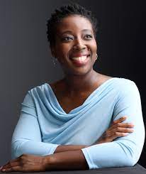 Headshot of Afua Bruce, an African American woman wearing a light blue top, arms crossed, staring out into the distance.