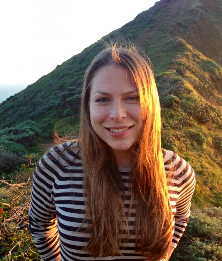 Headshot of Michelle Shevin, a white woman wearing a striped long sleeve shirt, smiling and posed on top of a grassy mountain.